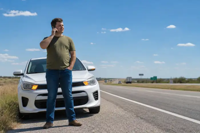 Driver standing beside a stalled car on the shoulder of a Dallas highway, calling for roadside assistance
